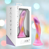 The Avant Sun's Out Dildo - Pink by Avant, featuring pink, yellow, and purple swirls and a suction cup base, stands next to its white and purple packaging. Made from platinum-cured silicone, it's set against a blurred pastel light background.